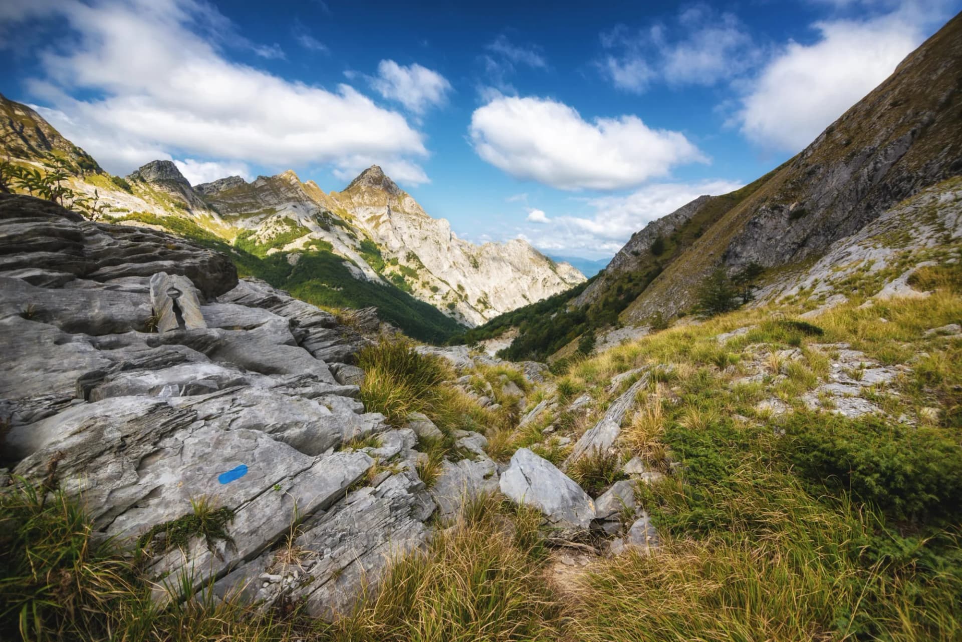 Hiking trail marker on rocky terrain leading into steep Apuan Alps valley under blue sky.