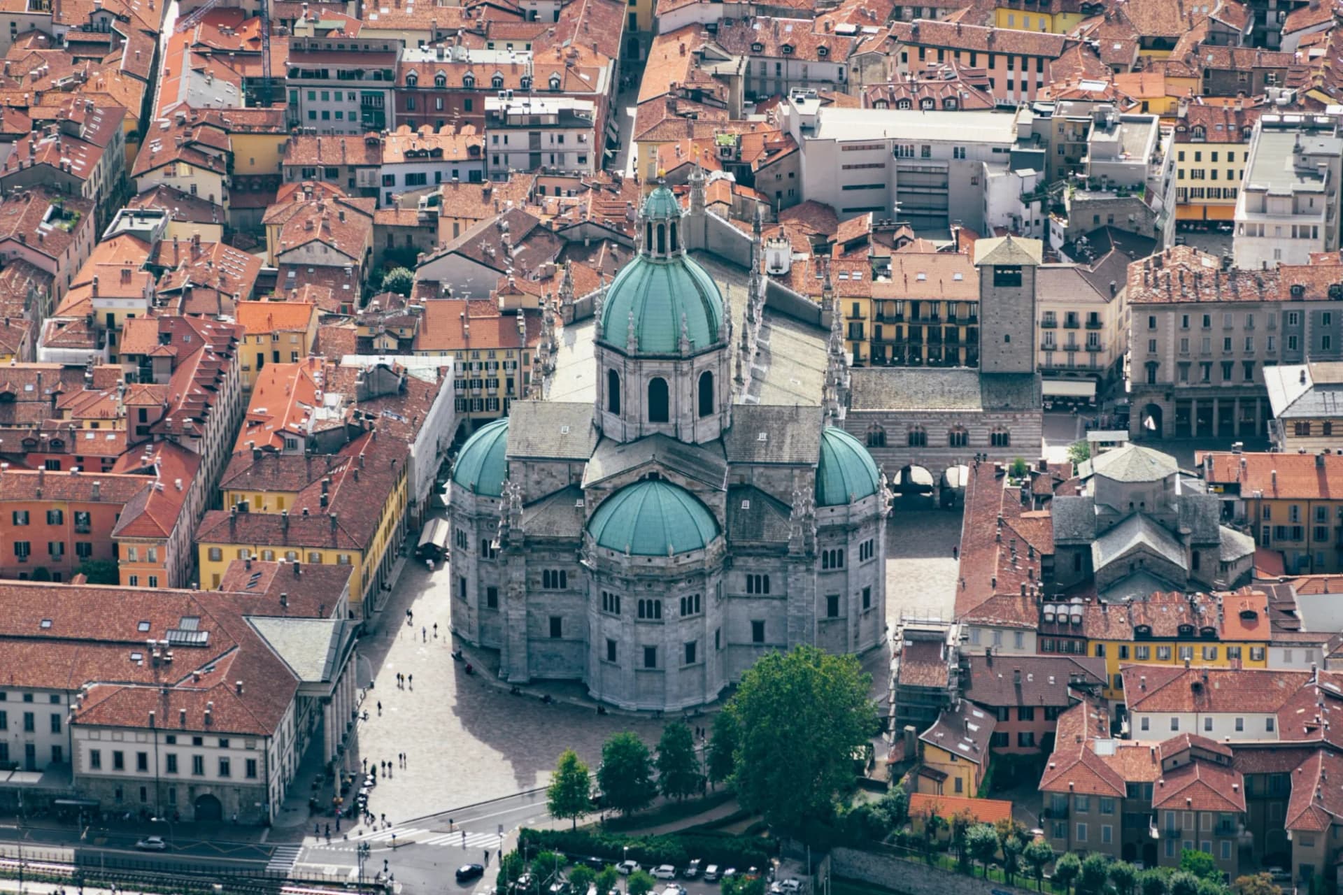 Cathedral with teal domes surrounded by dense terracotta-roofed buildings in a city square.