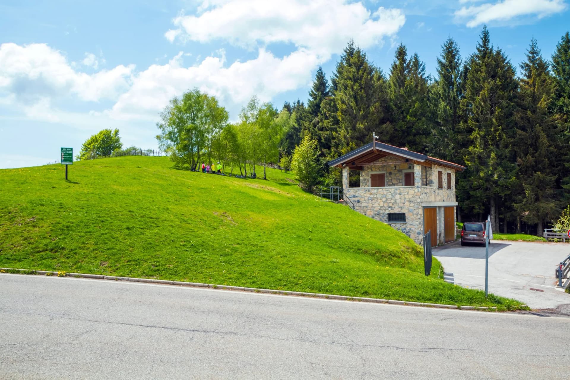 Stone building on a bright green hillside next to a dense pine forest under a blue sky.