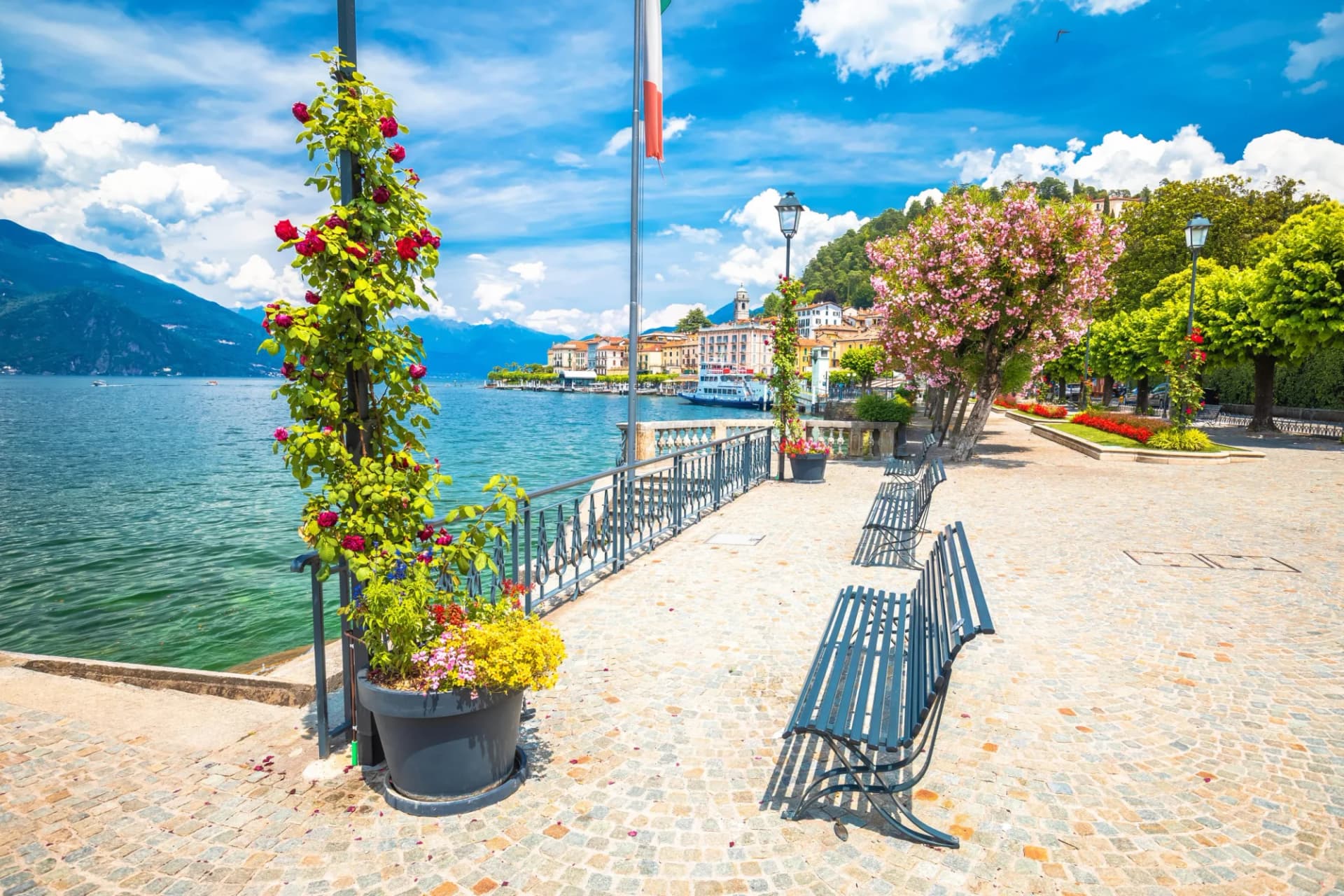 Lakeside promenade with cobblestones, benches, blooming roses, and Italian flag near mountains.