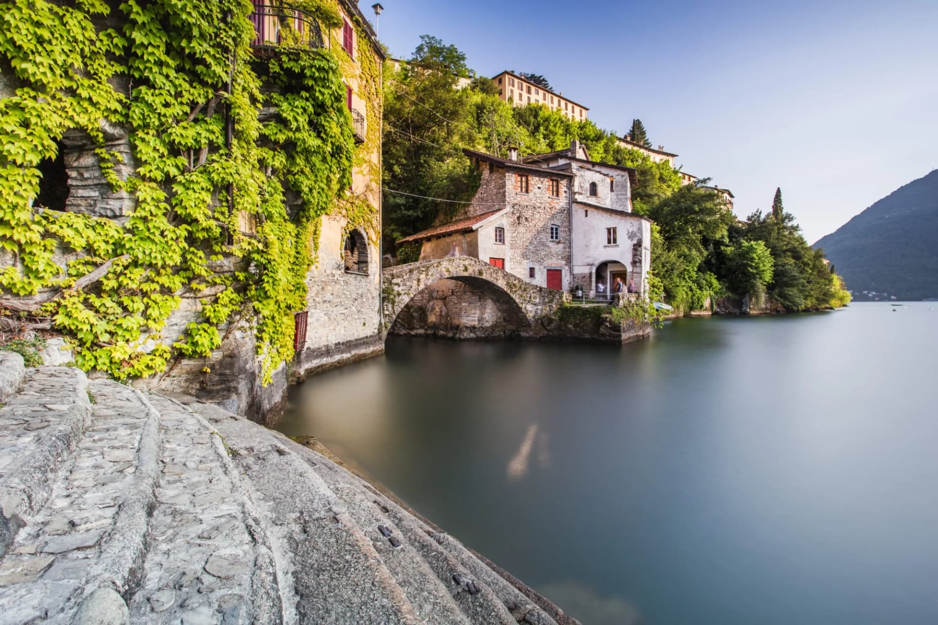 Stone steps leading to water by ivy-covered buildings and an arched bridge on Lake Como.