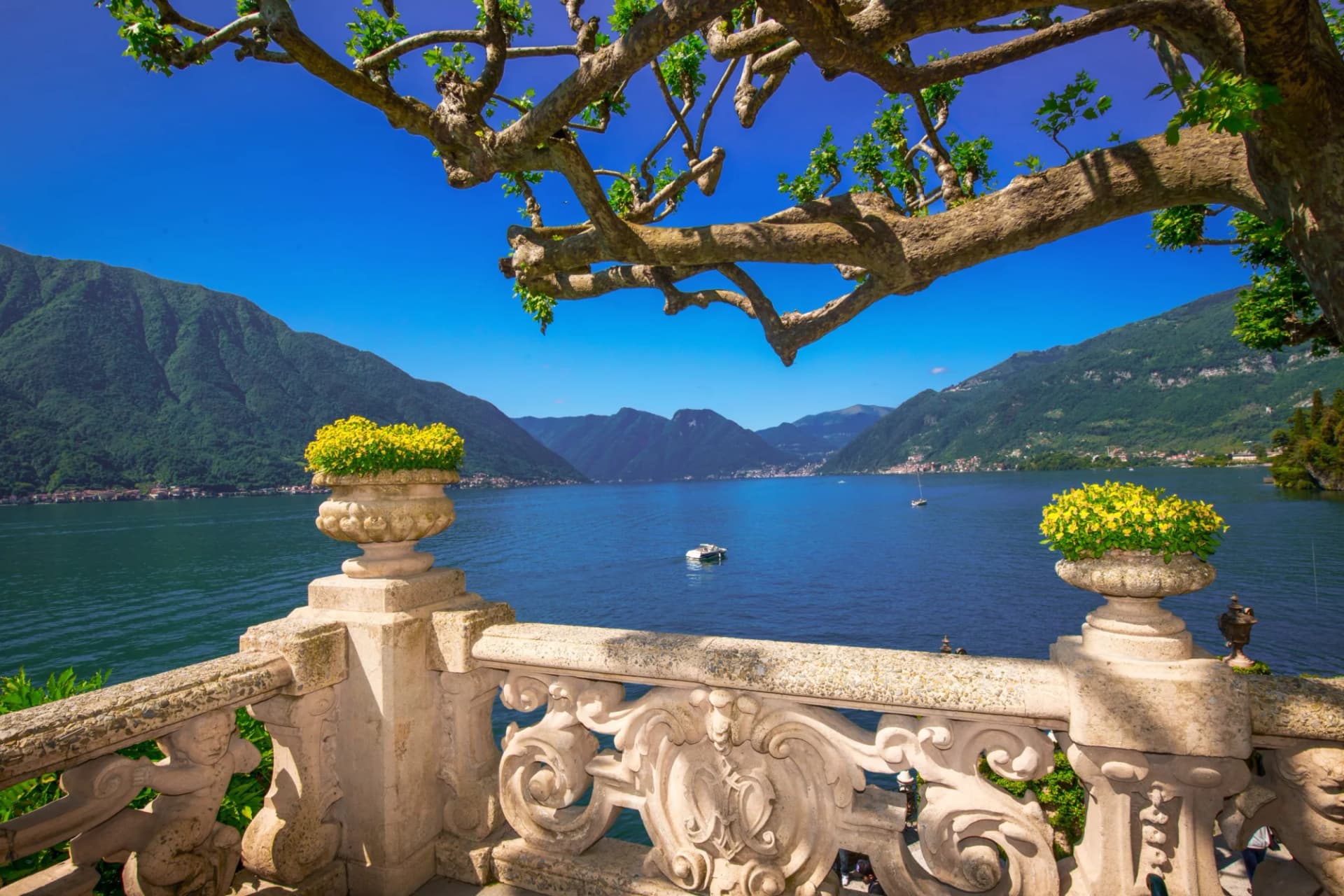 Ornate stone balcony overlooking Lake Como with mountains and boats under clear blue sky.