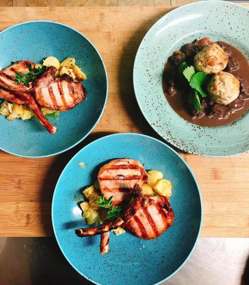 Three plates of grilled meat chops with potatoes and a plate of dumplings in brown sauce on a wooden surface.