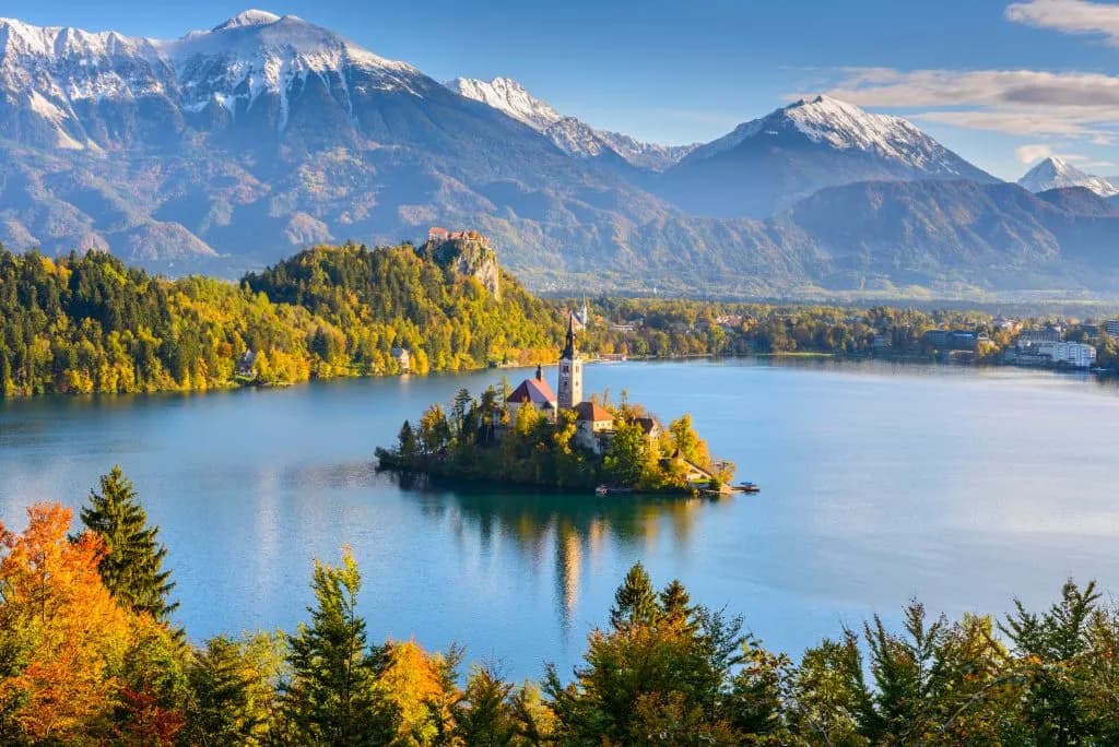 Panoramic view of Lake Bled island church with autumn trees and snow-capped mountains in Slovenia.