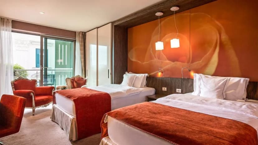 Twin beds in Hotel Kempinski Palace room with orange floral accent wall and red armchairs.