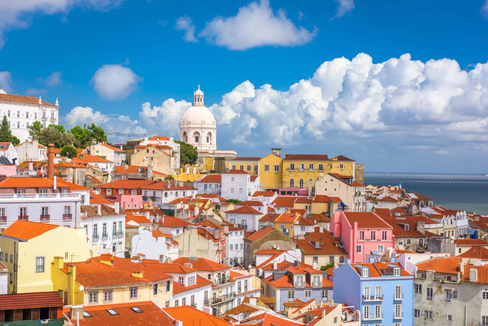 Lisbon old city skyline with colorful buildings, dome, and bridge over water under blue sky.
