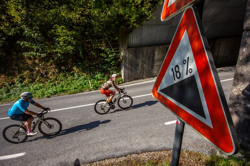 Cyclists ascending a steep road marked with an 18% grade sign near Wurzenpass.