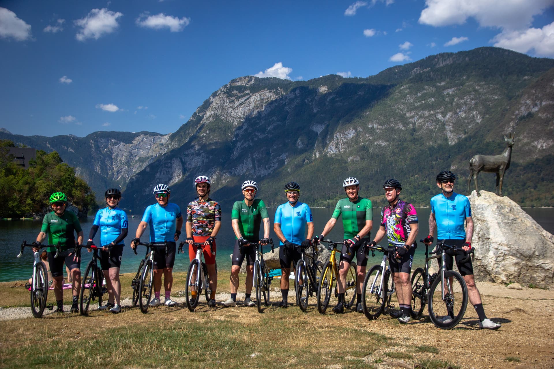 Cyclists pose with bikes by Bohinj Lake, with steep mountains and a bronze deer statue in the background.