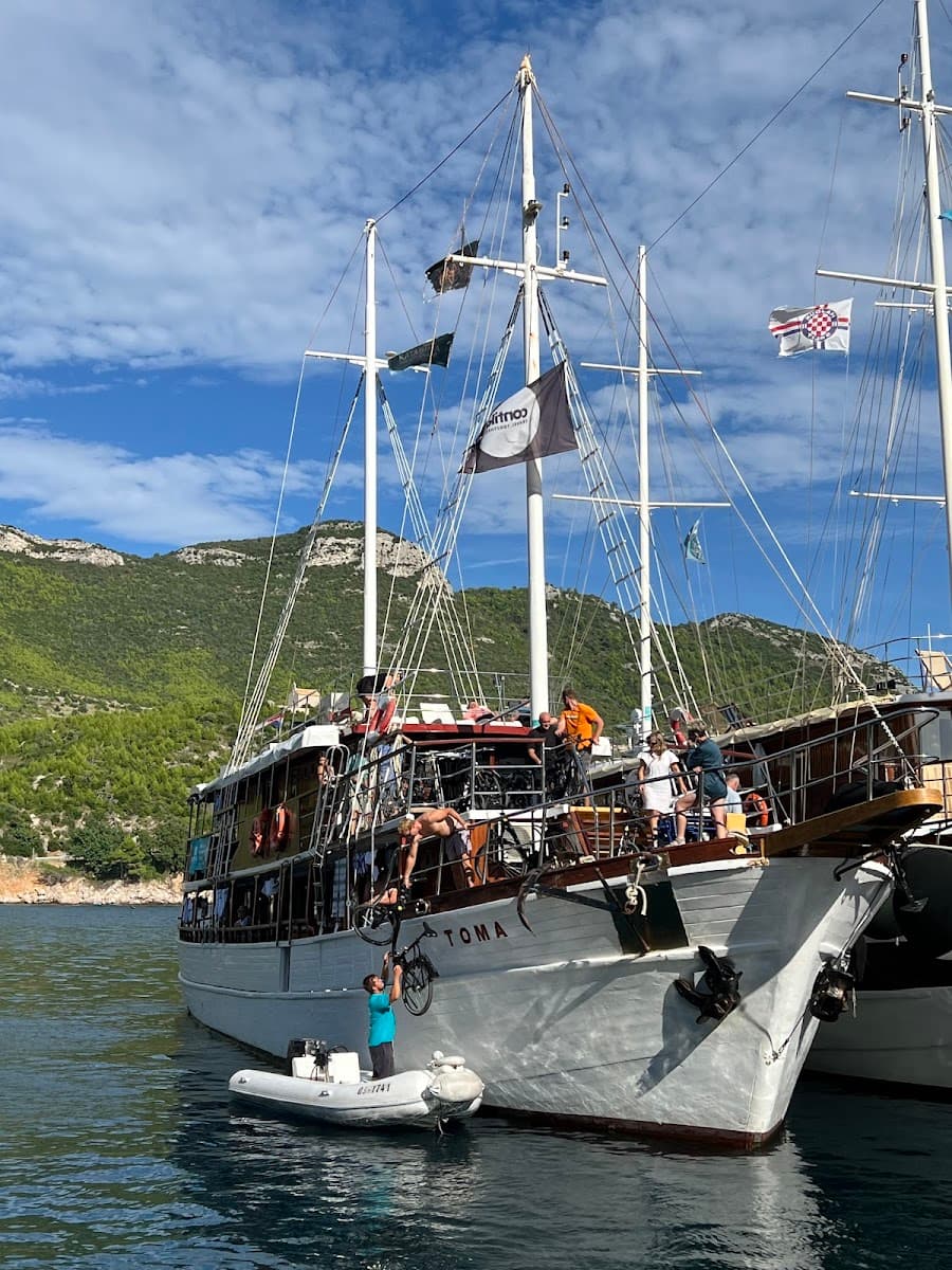 Large sailboat named Toma loading bicycles from a dinghy near a forested coastline.