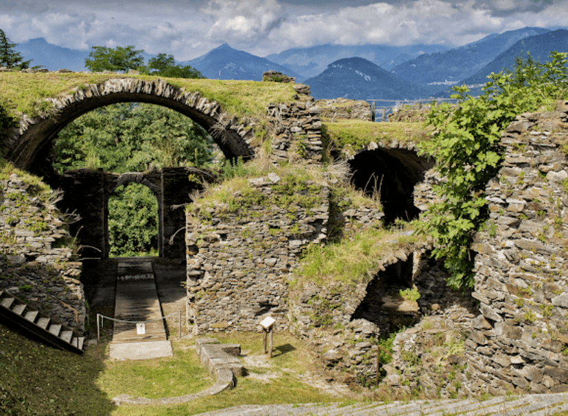 Stone ruins with arches overgrown with grass, overlooking mountains under a cloudy sky.