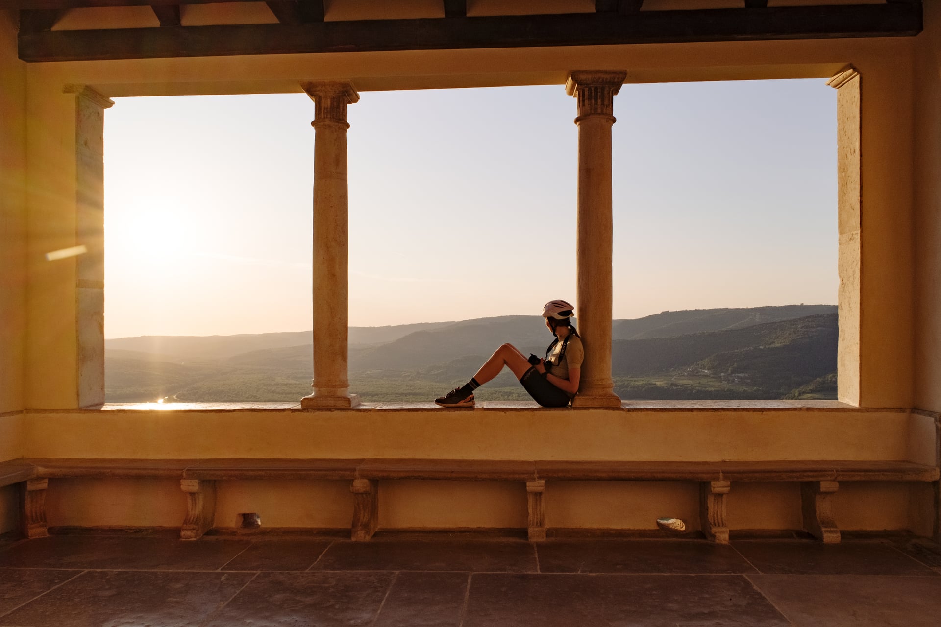 Cyclist resting inside stone loggia overlooking rolling green hills at sunset