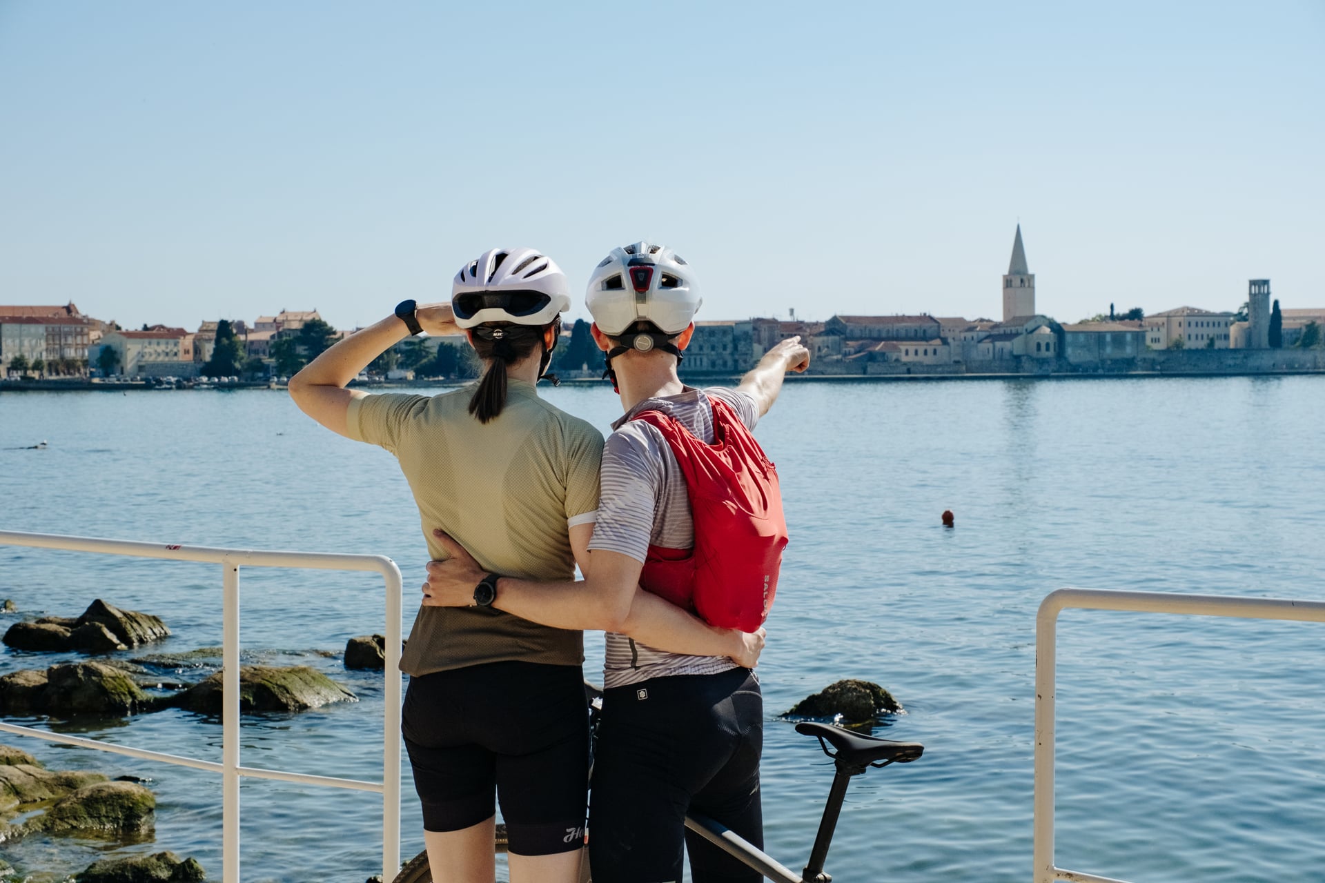 Cyclists with helmets look across the water toward a historic coastal town with a prominent bell tower.