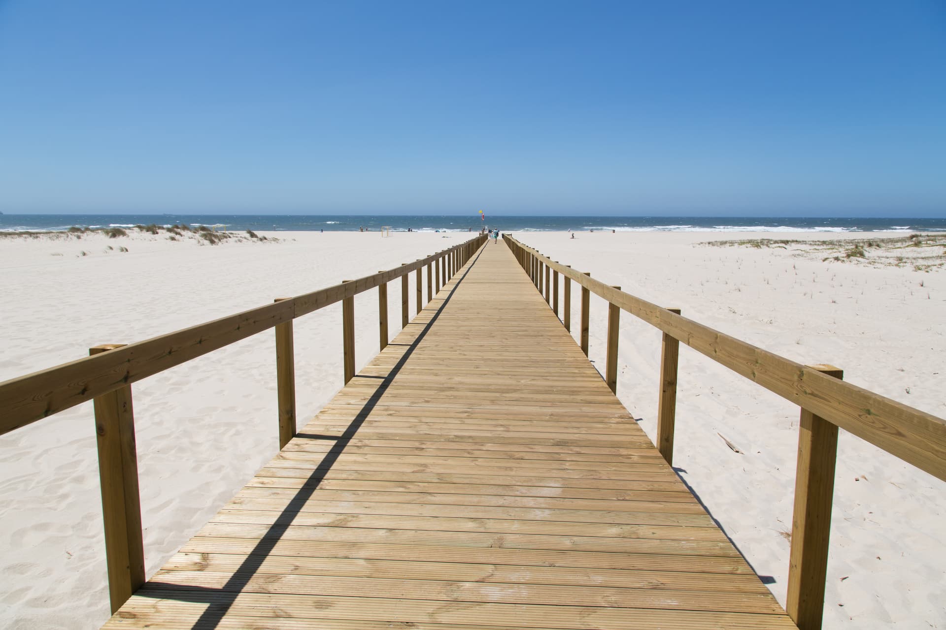 Wooden boardwalk leading across white sand dunes to the ocean under a clear blue sky in São Jacinto.