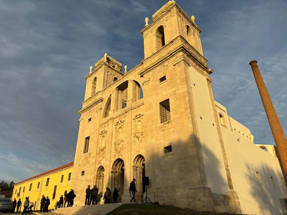 Seiça Monastery facade with twin towers, people gathering, and a tall brick chimney against a dramatic sky.