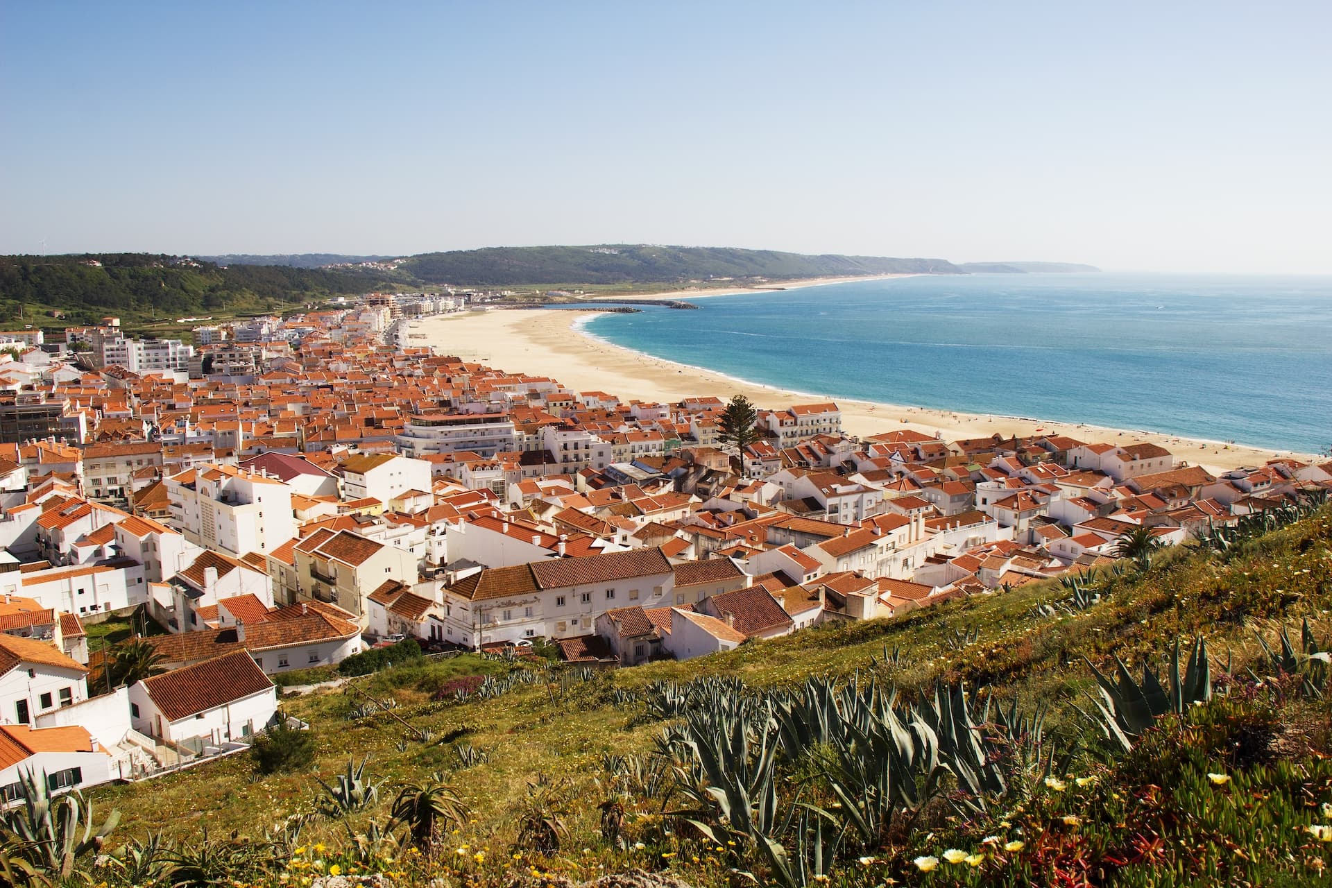 Coastal town of Nazare, Portugal with white buildings, terracotta roofs, and a long sandy beach meeting blue ocean.