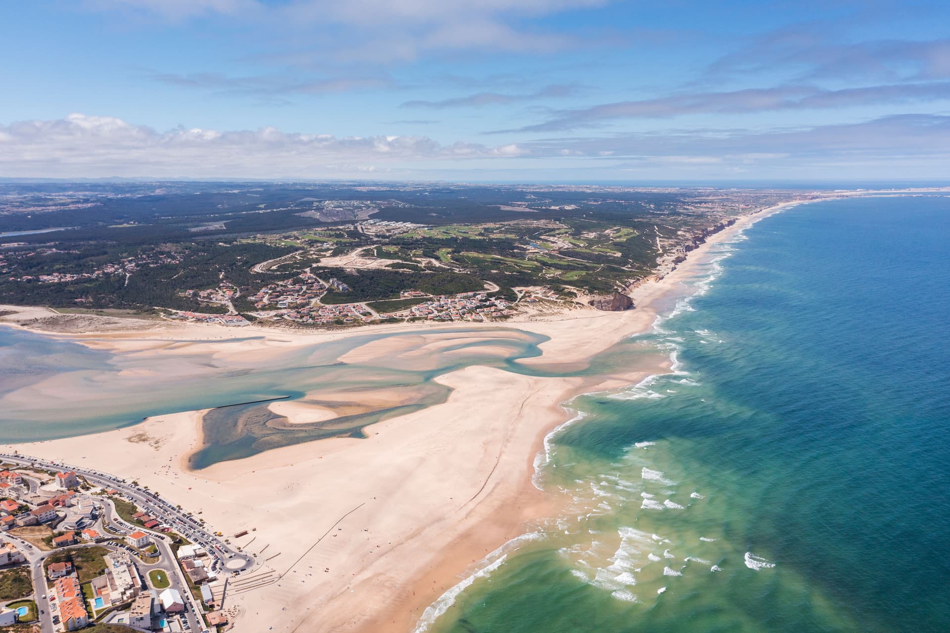 Aerial view of Foz do Arelho Beach with estuary, sandbars, town, and blue ocean waves.
