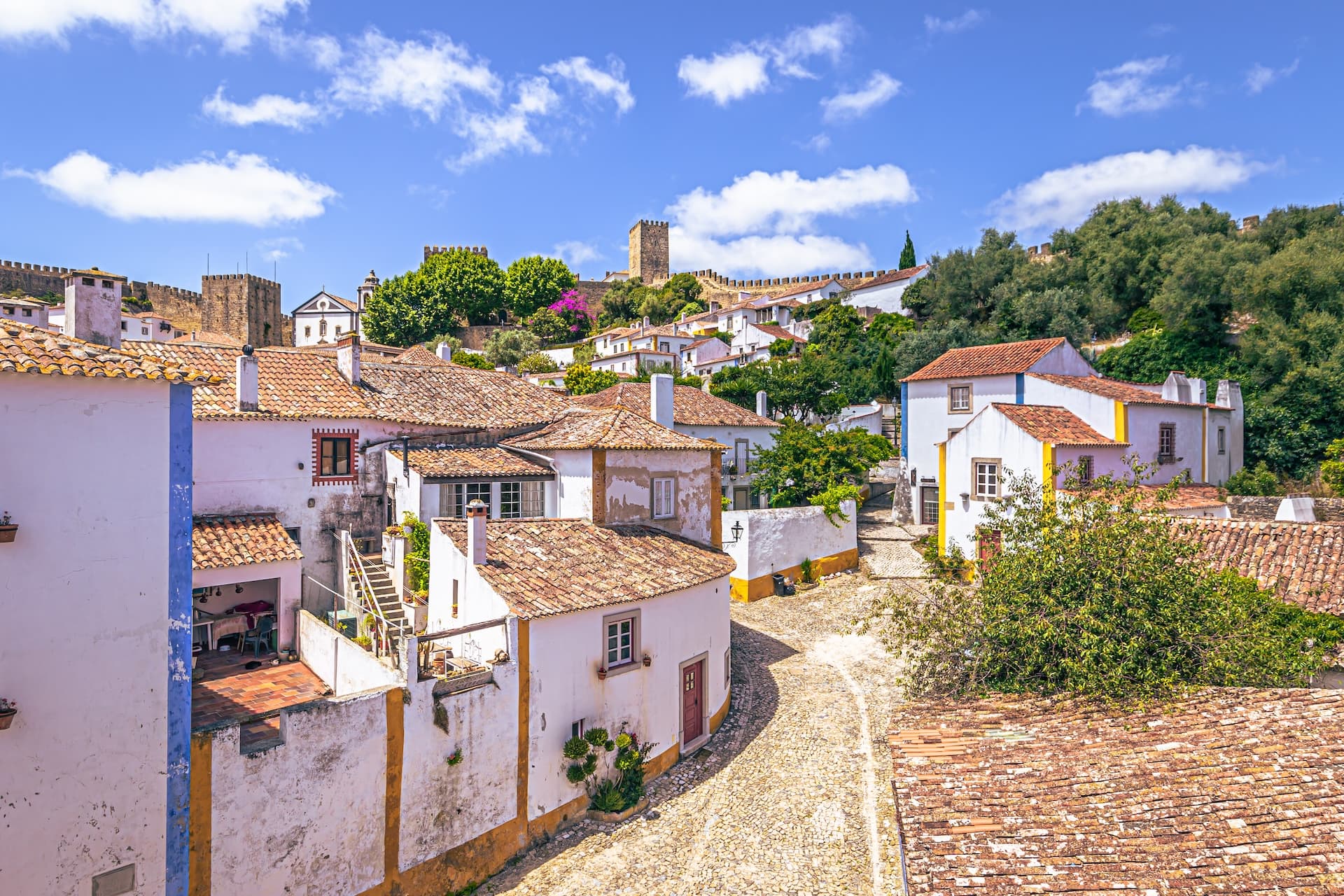 White houses with terracotta roofs climbing a hill toward a medieval castle wall in Obidos town.