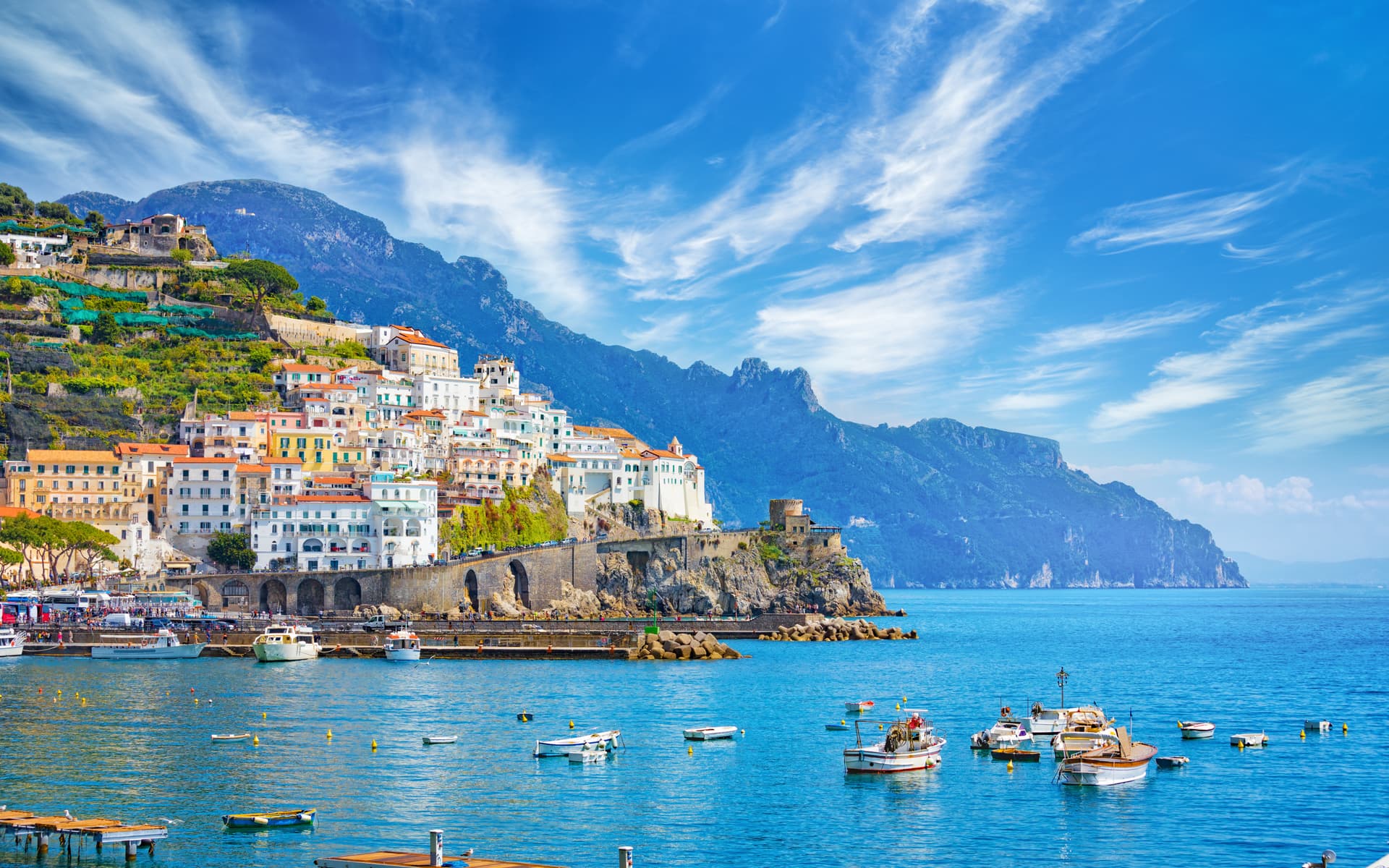 Coastal town with white buildings clinging to steep green mountains above bright blue Mediterranean water.