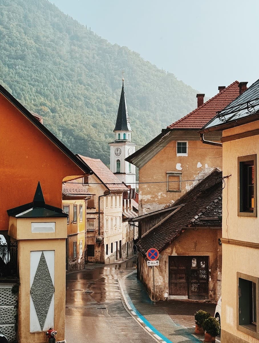Wet, narrow street in a European town leading to a church spire, backed by a forested mountain.