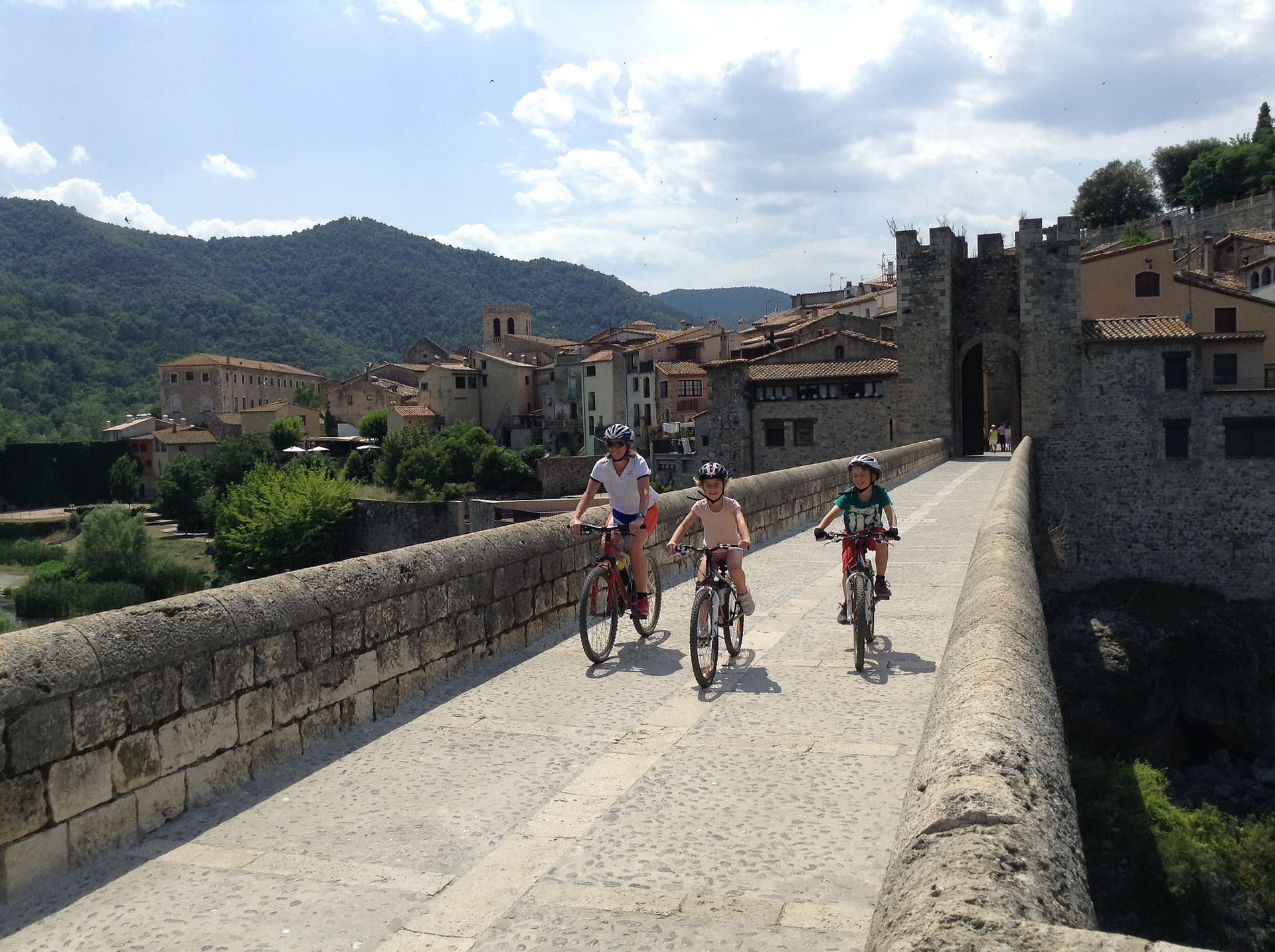 Family cycling across a historic stone bridge toward a medieval town in Girona.