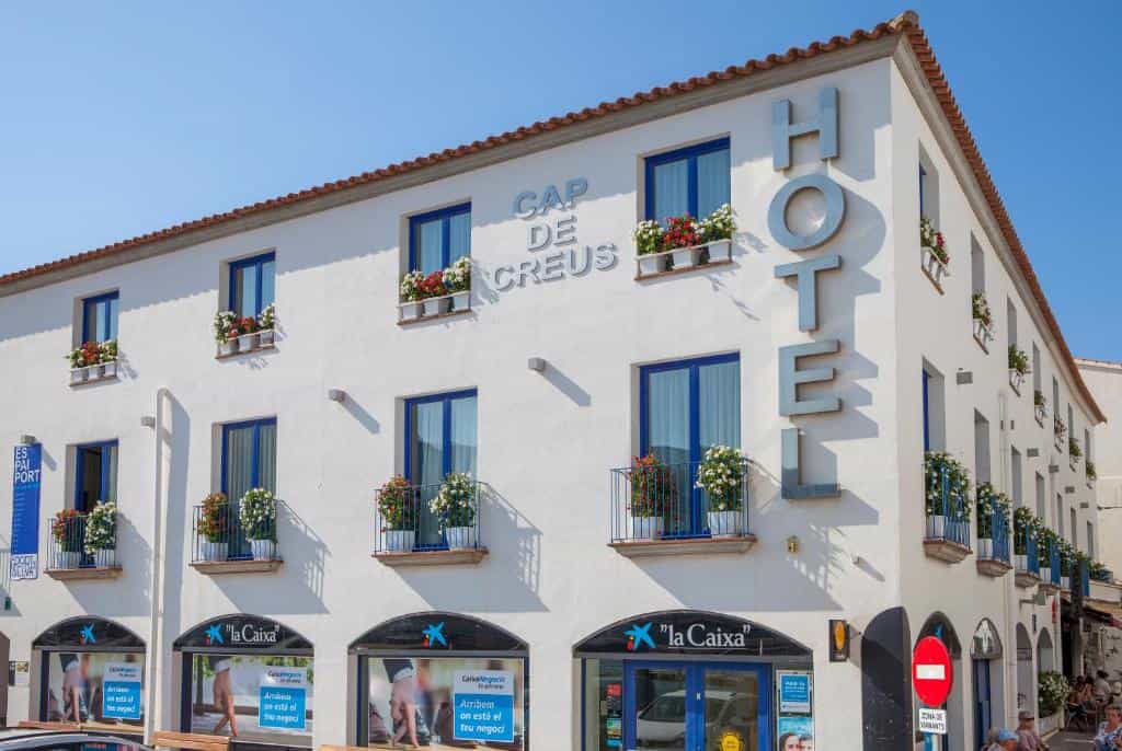 Hotel Cap de Creus building with white facade, blue window frames, and flower-filled balconies.