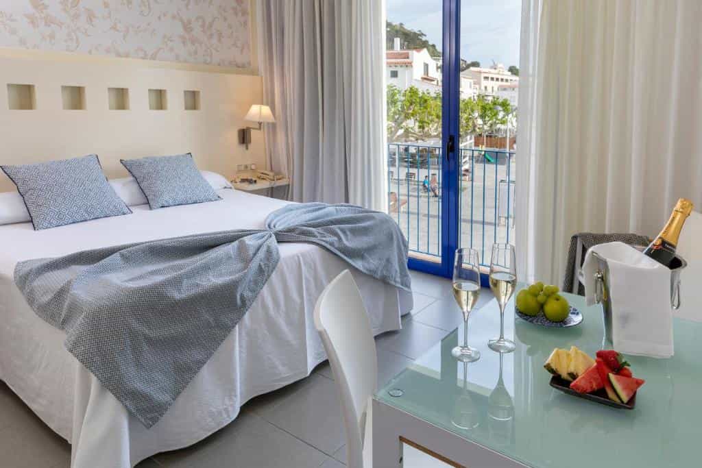 Hotel room with double bed, champagne, and fruit, overlooking a coastal town square.
