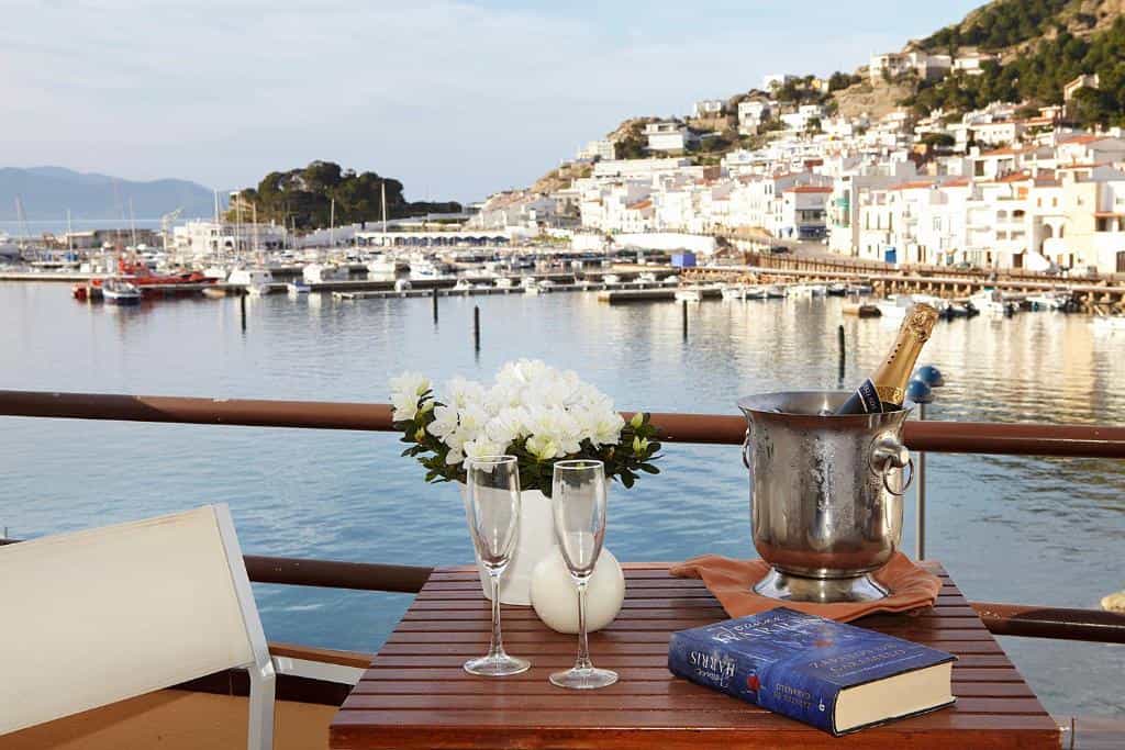 Champagne in ice bucket with glasses and book on a balcony overlooking a coastal harbor village.