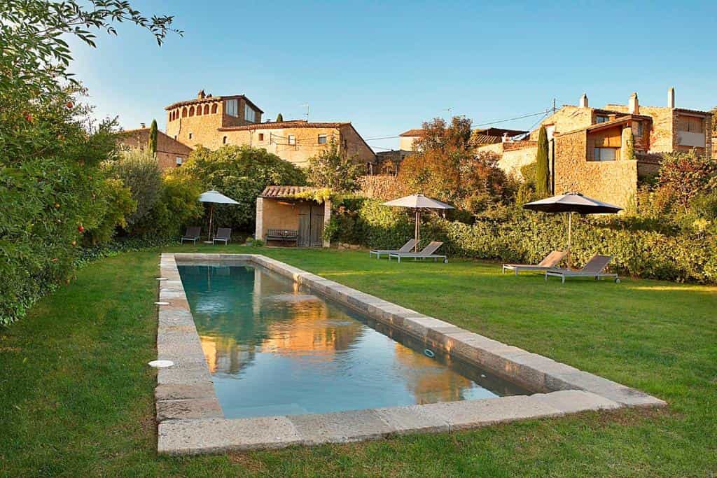 Swimming pool in a garden with lounge chairs and umbrellas in front of stone village buildings.