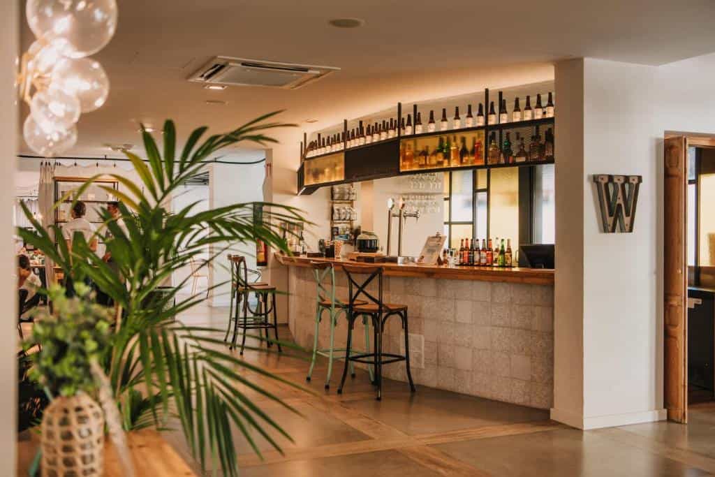 Bar counter with stools, shelves of bottles, and indoor plants in a restaurant setting.
