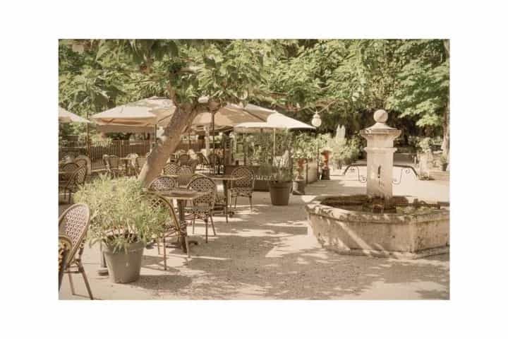 Outdoor cafe seating with tables, chairs, and umbrellas next to a stone fountain in a shaded courtyard.