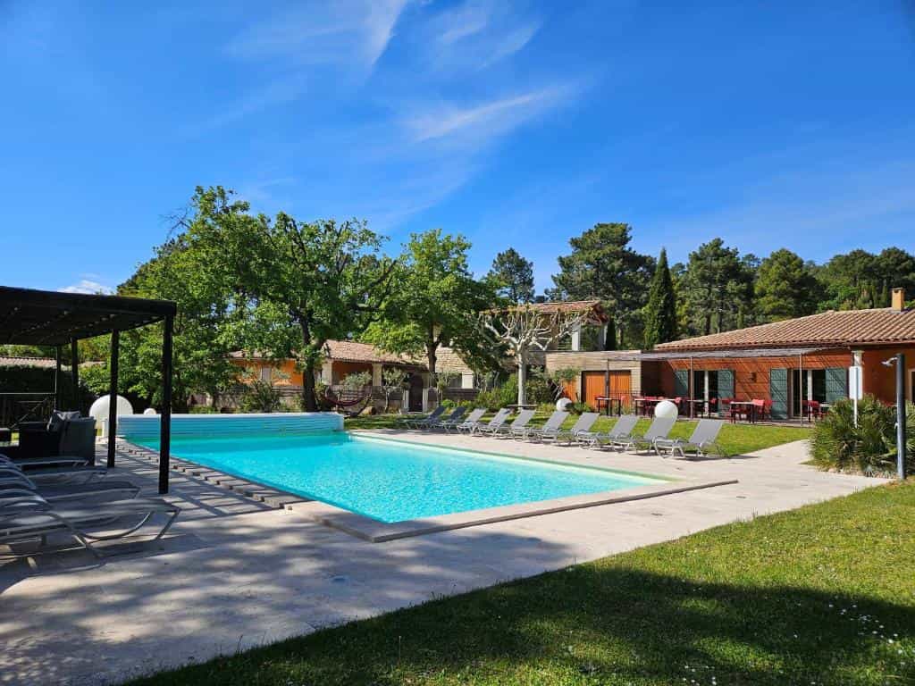 Swimming pool with lounge chairs on a sunny patio surrounded by trees and Mediterranean-style buildings.