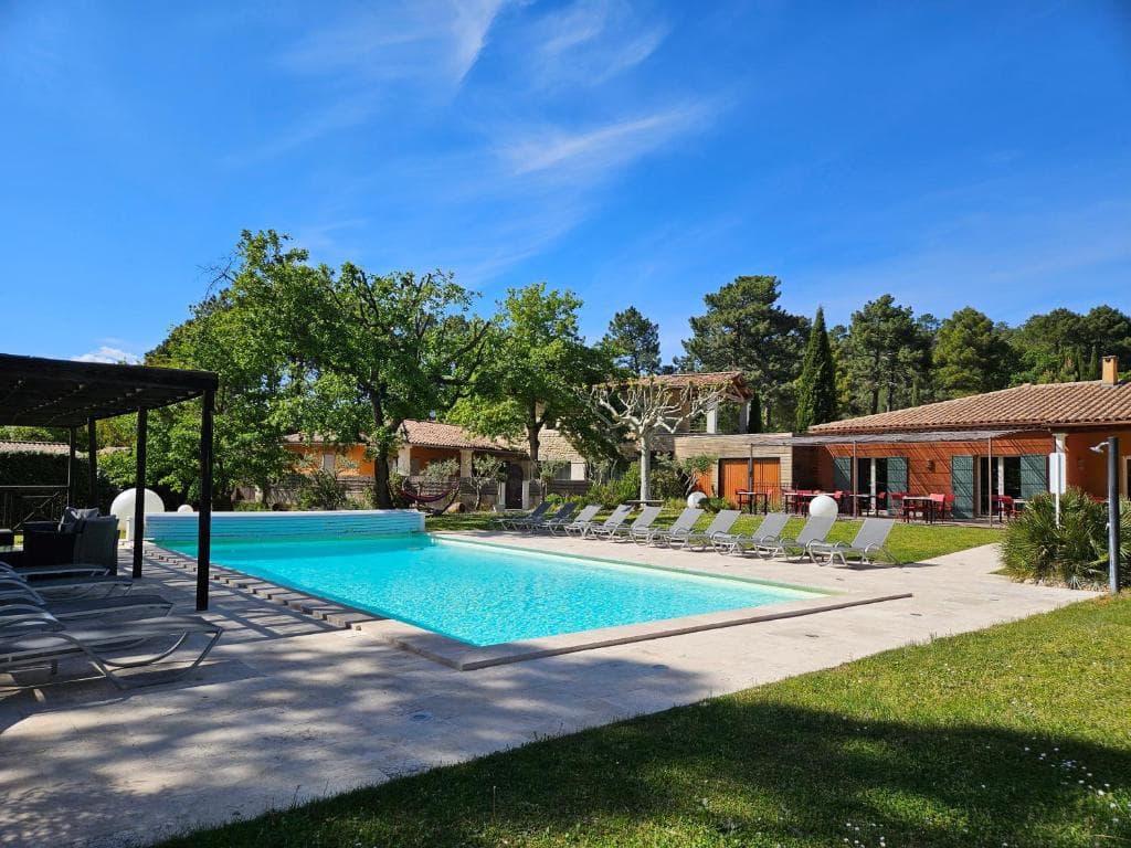 Swimming pool with lounge chairs on a sunny patio surrounded by trees and Mediterranean-style buildings.