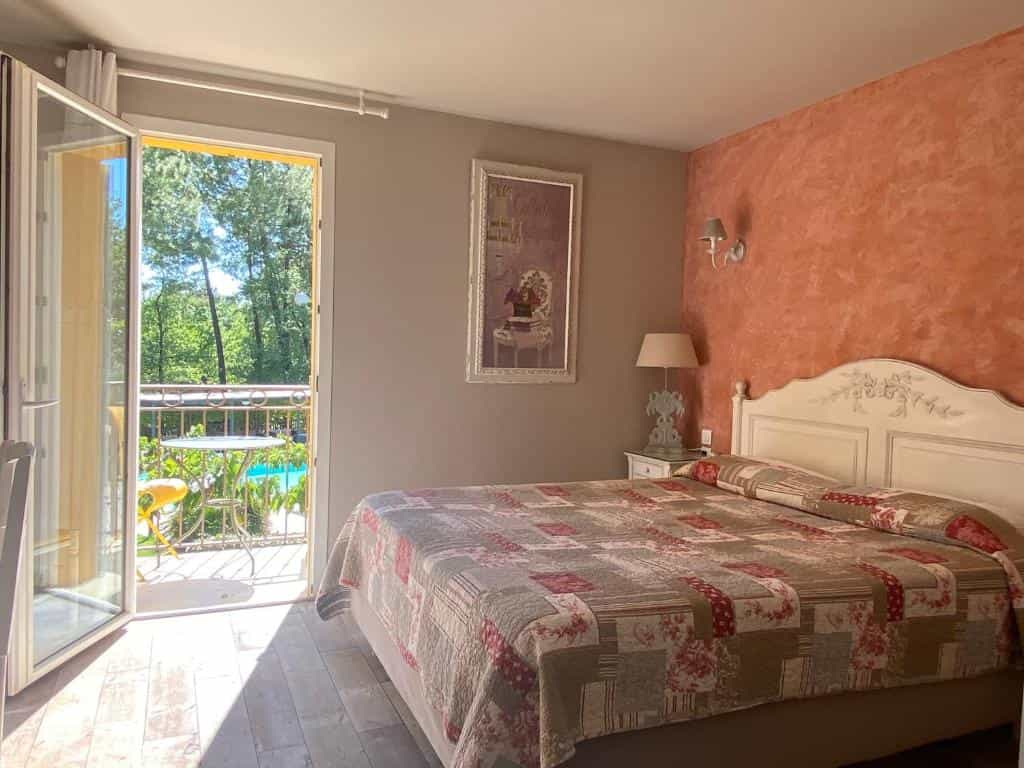 Hotel bedroom with patchwork quilt bed, terracotta accent wall, and balcony overlooking trees and a swimming pool.