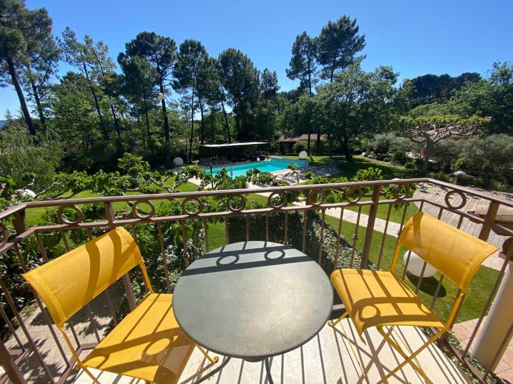 Yellow chairs and a small table on a balcony overlooking a swimming pool in a garden with tall pine trees.