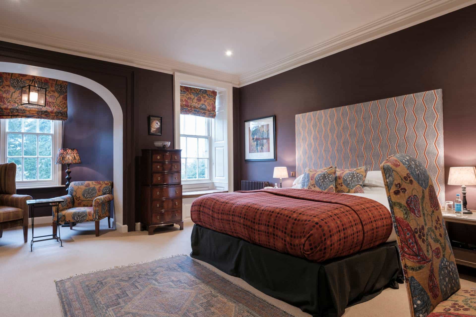 Hotel bedroom with dark purple walls, patterned headboard, and seating area by the window.