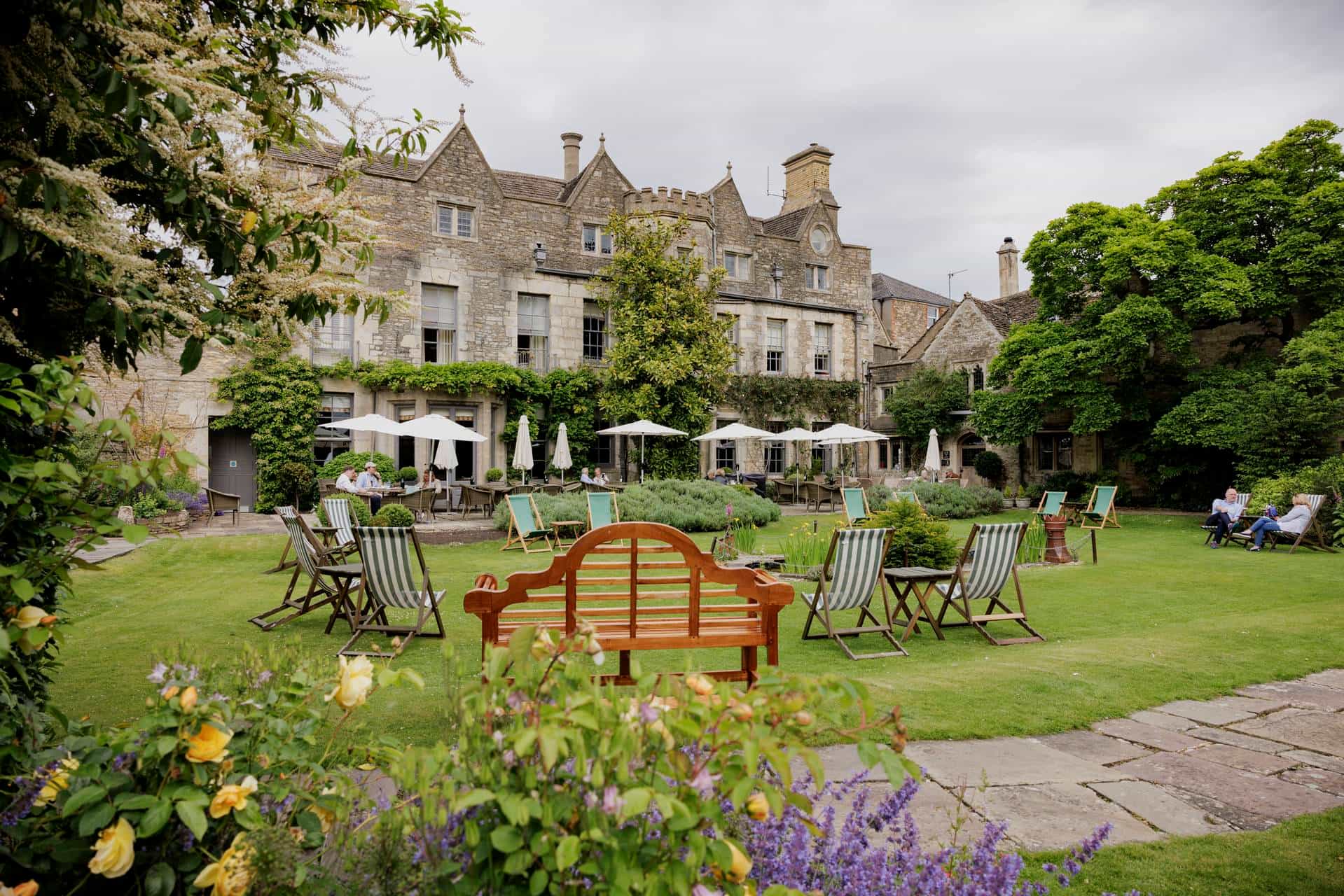 People relaxing in the garden of The Close in the Cotswolds with stone buildings, lawn chairs, and blooming flowers.