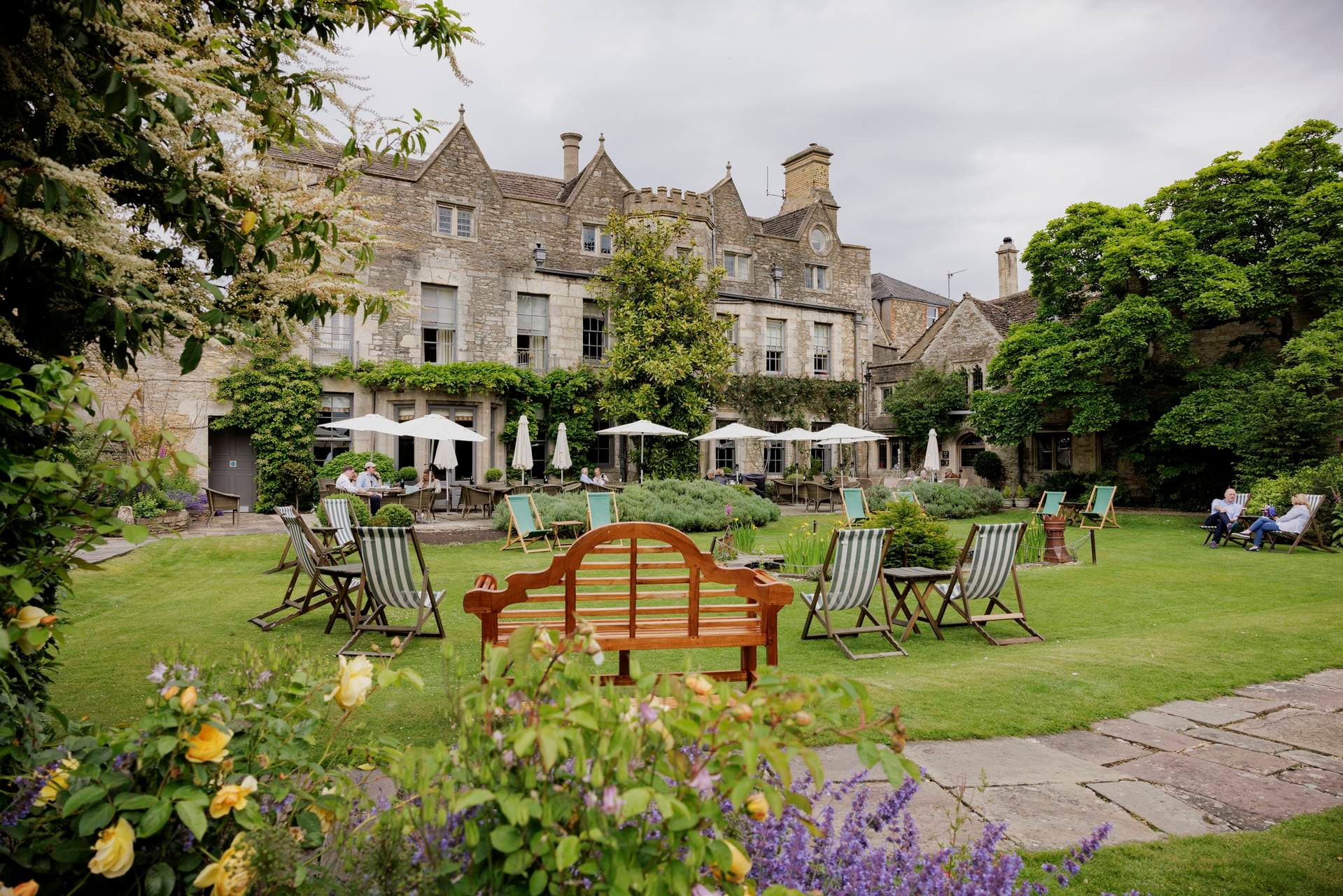 People relaxing in the garden of The Close in the Cotswolds with stone buildings, lawn chairs, and blooming flowers.