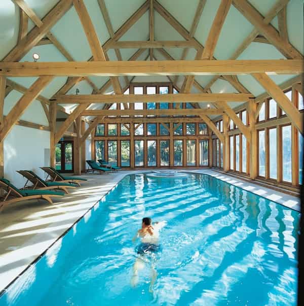 Person swimming in an indoor pool with timber frame architecture and lounge chairs.