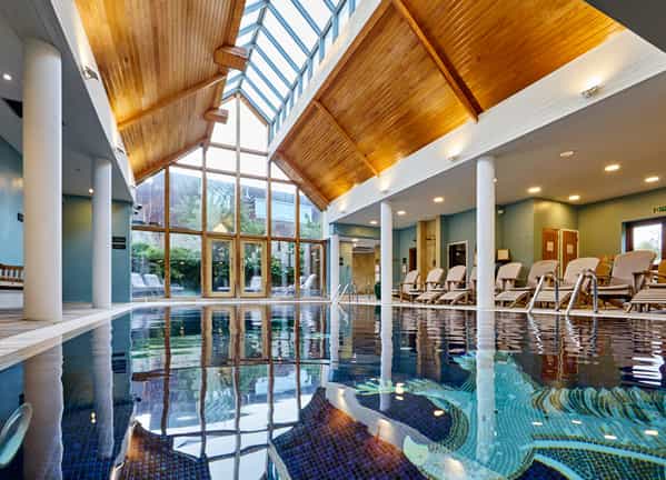 Indoor swimming pool with a glass-paneled roof, wooden vaulted ceiling, and lounge chairs on the deck.