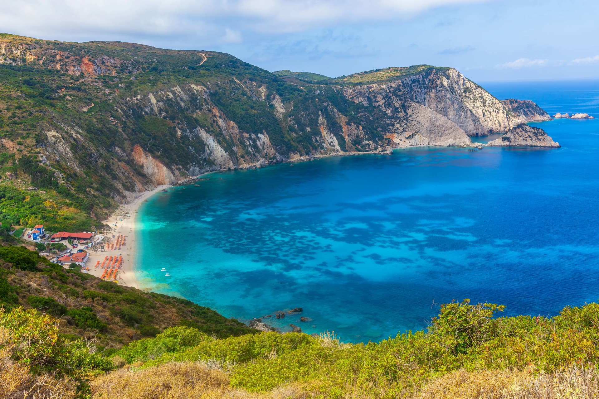 Petani Beach, Lixouri in Summer, on the Ionian island of Kefalonia, Greece, with orange umbrellas, sunbeds and clear blue sky and high mountains. Space for copy, Horizontal.