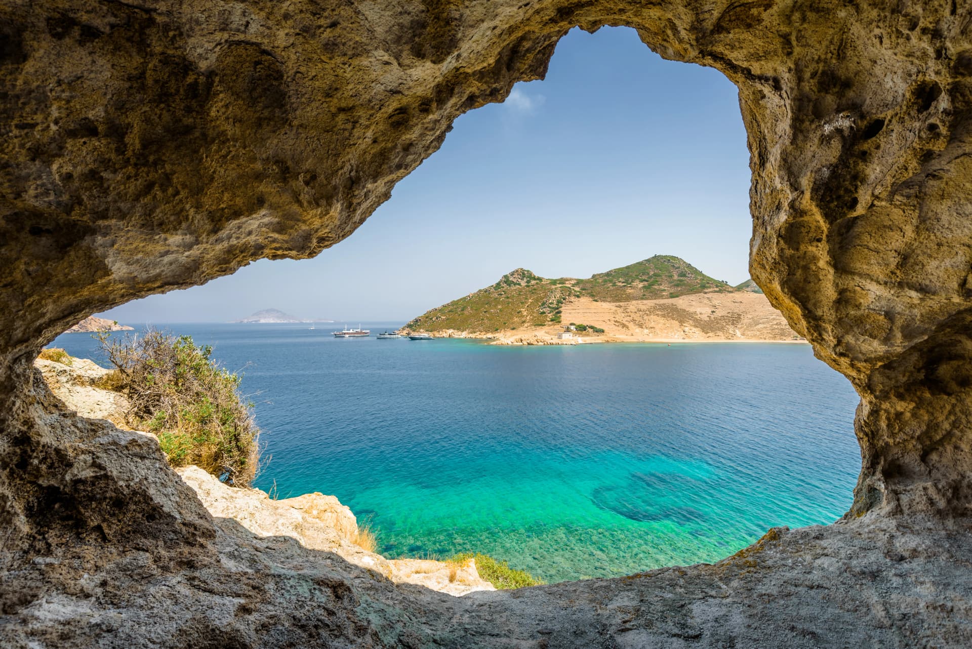 Beautiful sunny view through a rocky cave to the greek blue sea with crystal clear water from an a hill with boats cruising fishing surrounded by mountains, Patmos Island, Kos, Dodecanese/ Greece