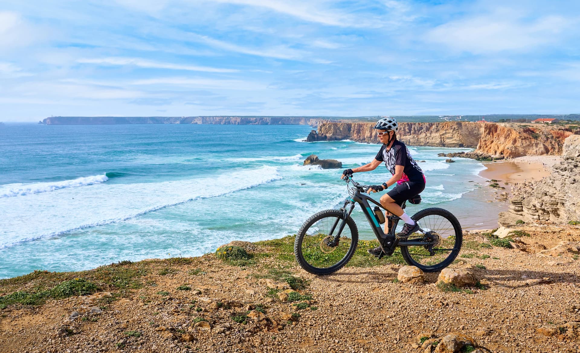 happy active senior woman cycling at the the rock cliffs and lighthouse of Cabo Sao Vicente, the south-western spit of Europe at the atlantic coast of Algarve, Portugal,