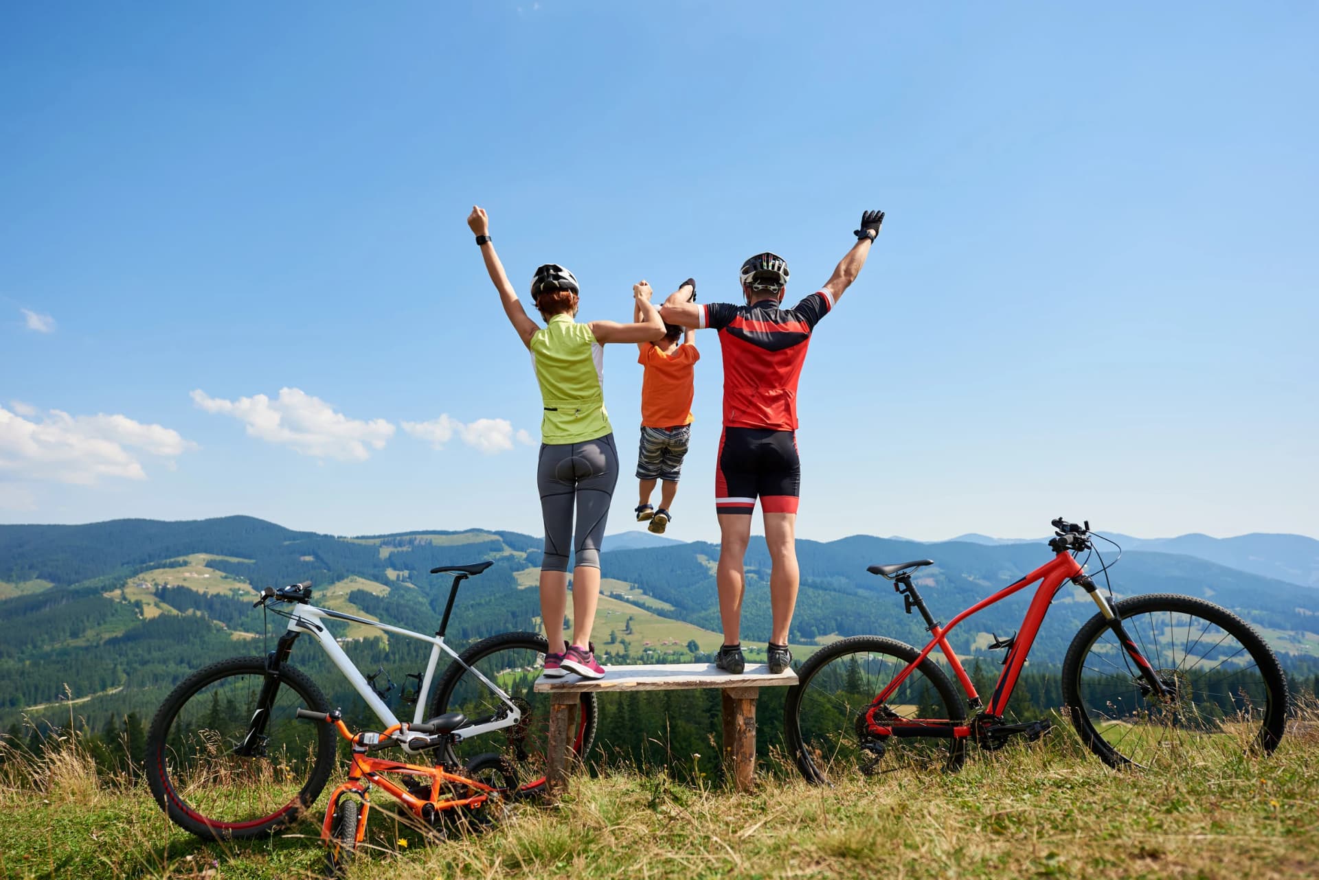 Rear view of family bikers, mom, dad standing on wooden bench with raised hands, holding child in the air, resting after cycling bicycles in mountains. Active lifestyle and happy relations concept.