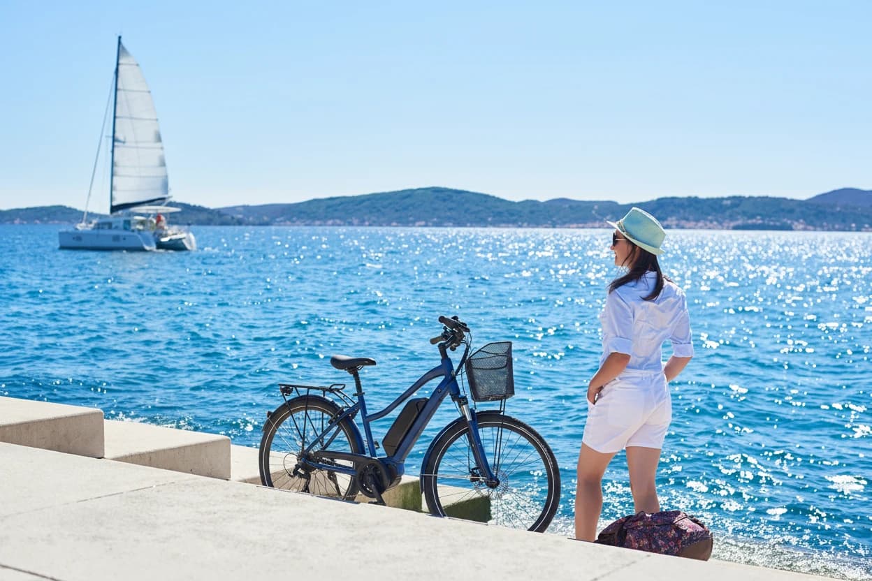Back view of slim tourist woman in white clothing and sunglasses resting at bicycle and backpack on paved stone sidewalk stairs on clear blue sea water, sailing cruse yacht and mountains background.