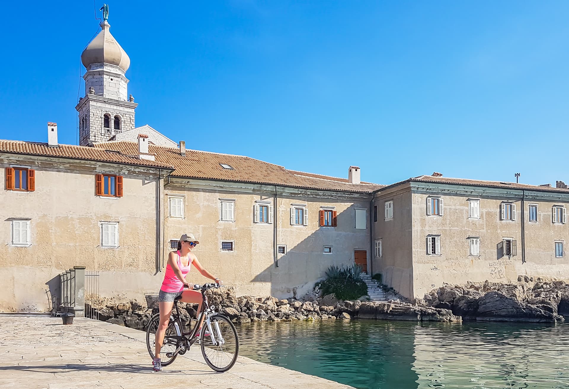Woman in pink tank top standing on a bicycle by the sea in the historic city center of Krk, Croatia.