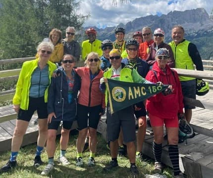 Group of cyclists holding an AMC 150 pennant in front of mountain peaks